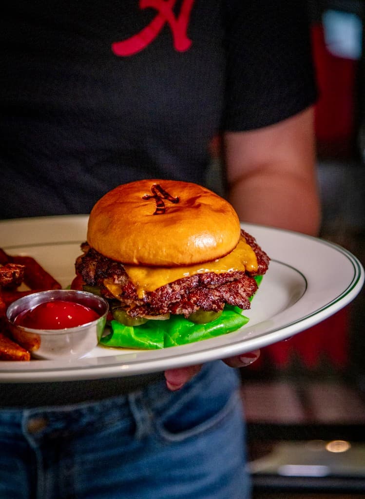 Server in an Archie's tee holding a plated double smashburger with melted cheddar, lettuce, and pickles on a branded bun, with fries and ketchup