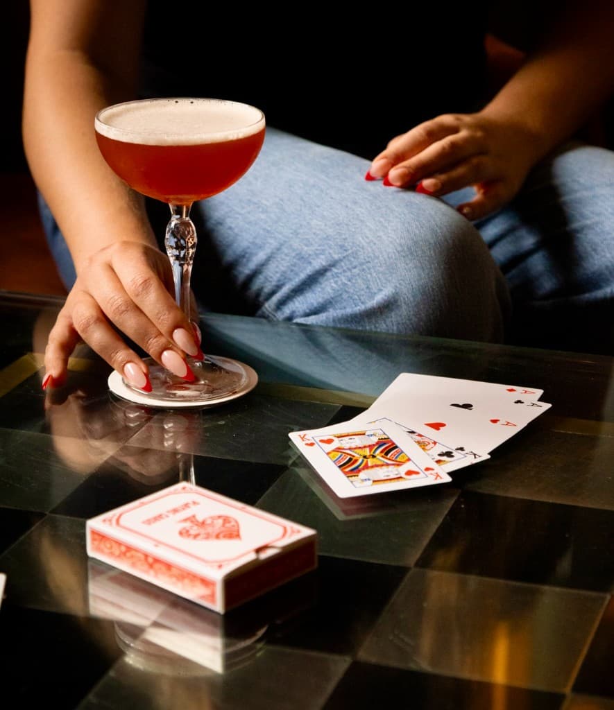 Hand with red-tipped nails resting a foamy amber cocktail on a coaster next to scattered aces, kings, and a deck of cards on a checkered table