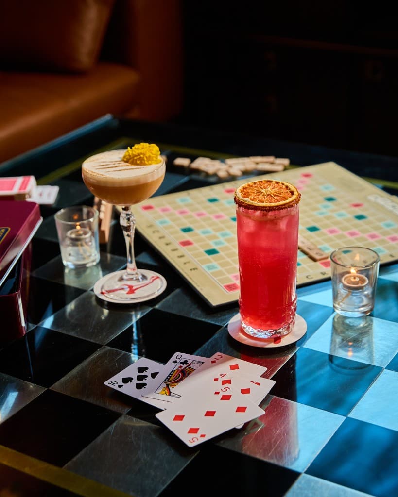 A foamy coupe cocktail and a bright pink highball with a dried citrus wheel on a checkered games table, beside a Scrabble board and playing cards lit by candles