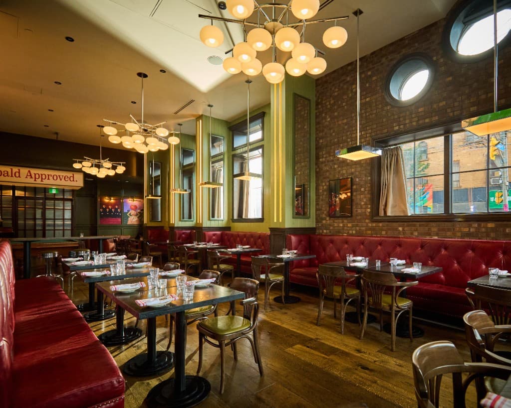 Archie's dining room with tufted red leather banquettes, wood chairs, globe chandeliers, green panelled walls, and round porthole windows on a brick wall