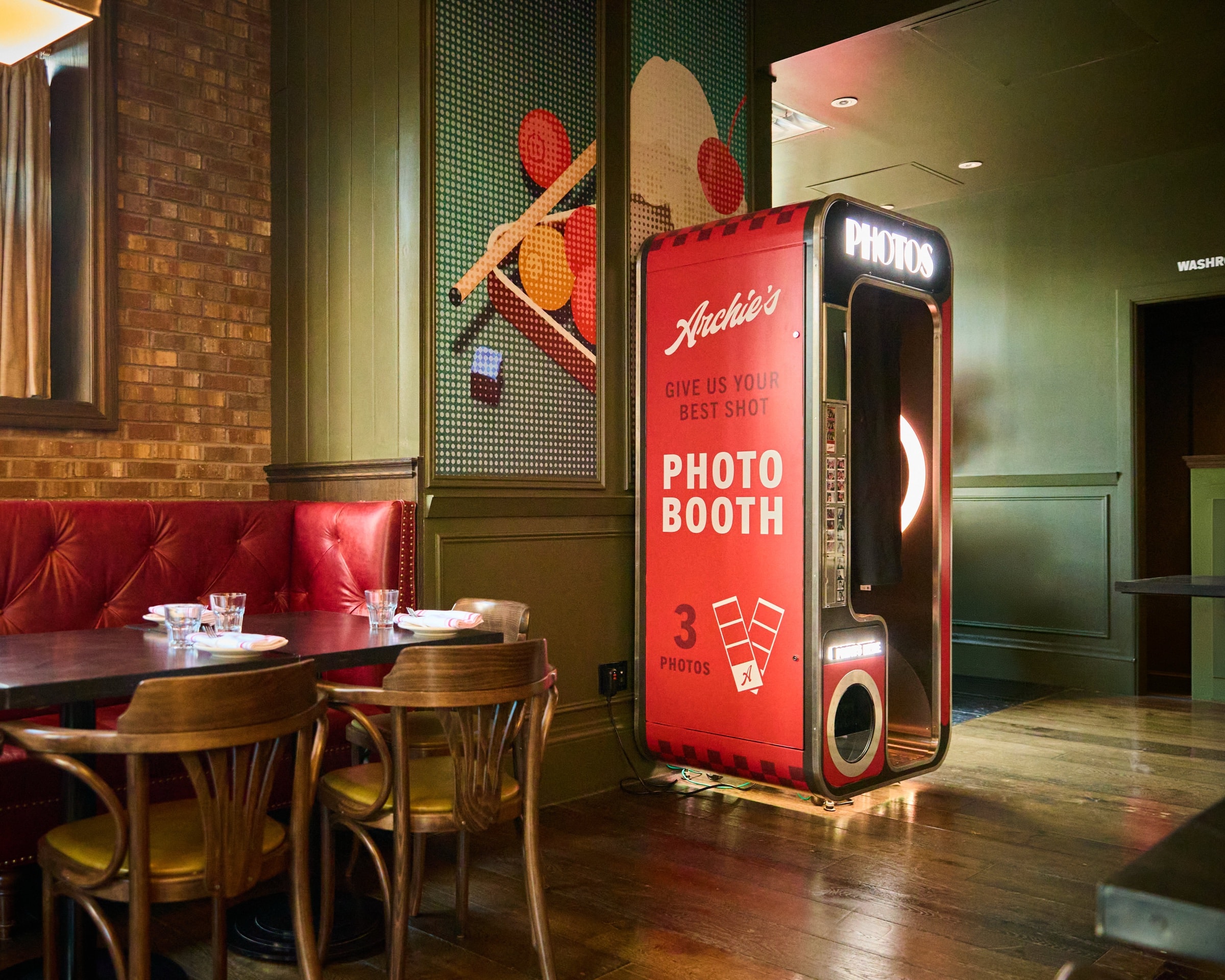 Archie's red photo booth beside set dining tables, red banquettes, brick, and green panelled walls