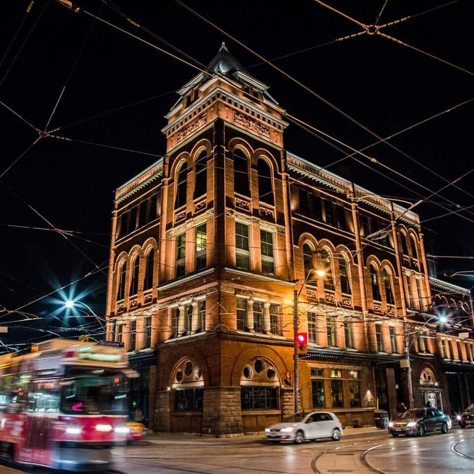The Broadview Hotel’s red-brick Romanesque corner building at night, warmly lit, with a streetcar passing on Broadview Avenue