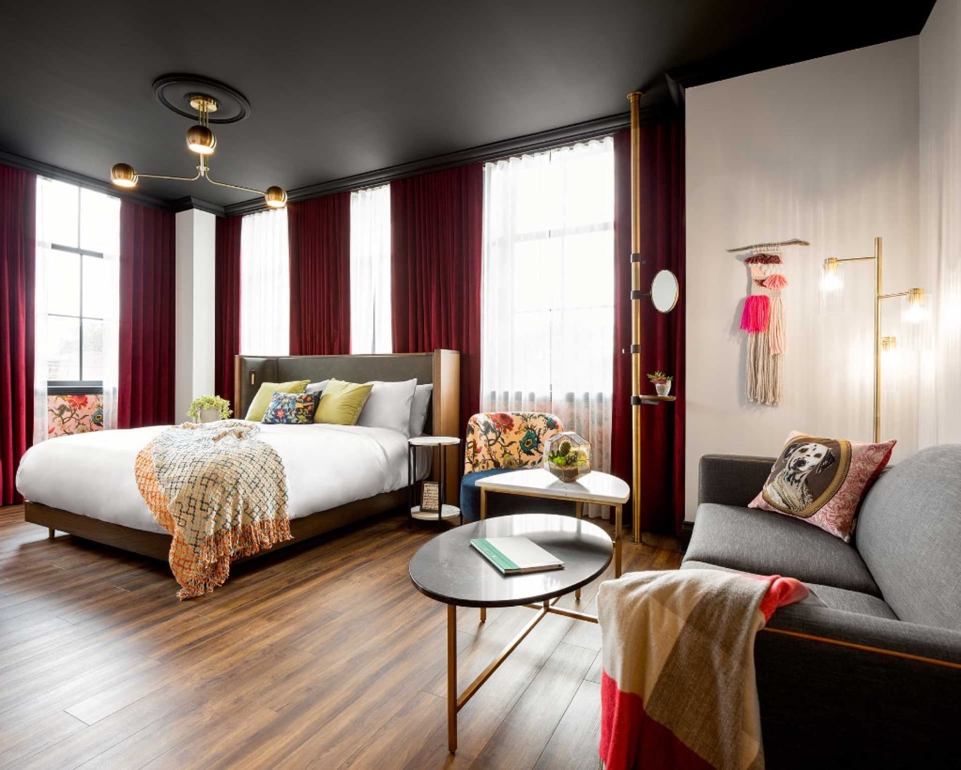 Boutique hotel king room with burgundy drapes, white bed linens, grey sofa with patterned pillows, floral armchair, marble coffee tables, and black ceiling with brass chandelier