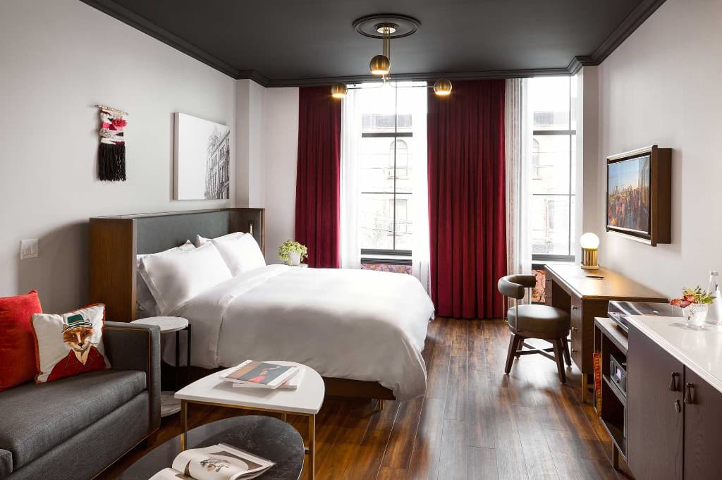 Boutique hotel king room with black ceiling and brass chandelier, white bed, grey sofa, burgundy floor-length drapes, marble coffee tables, and a wood desk with chair