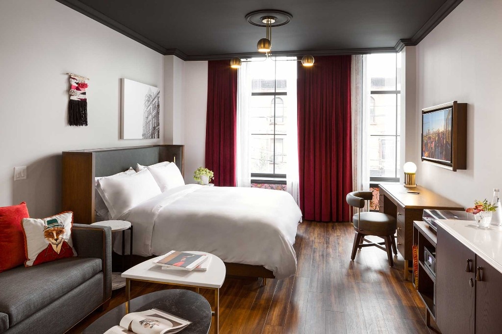 Boutique hotel king room with black ceiling and brass chandelier, white bed, grey sofa, burgundy floor-length drapes, marble coffee tables, and a wood desk with chair