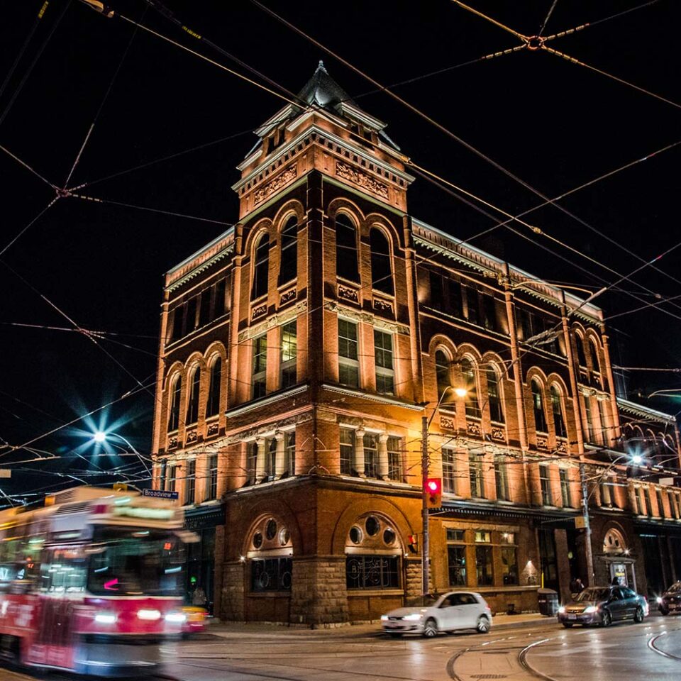 The Broadview Hotel’s red-brick Romanesque corner building at night, warmly lit, with a streetcar passing on Broadview Avenue
