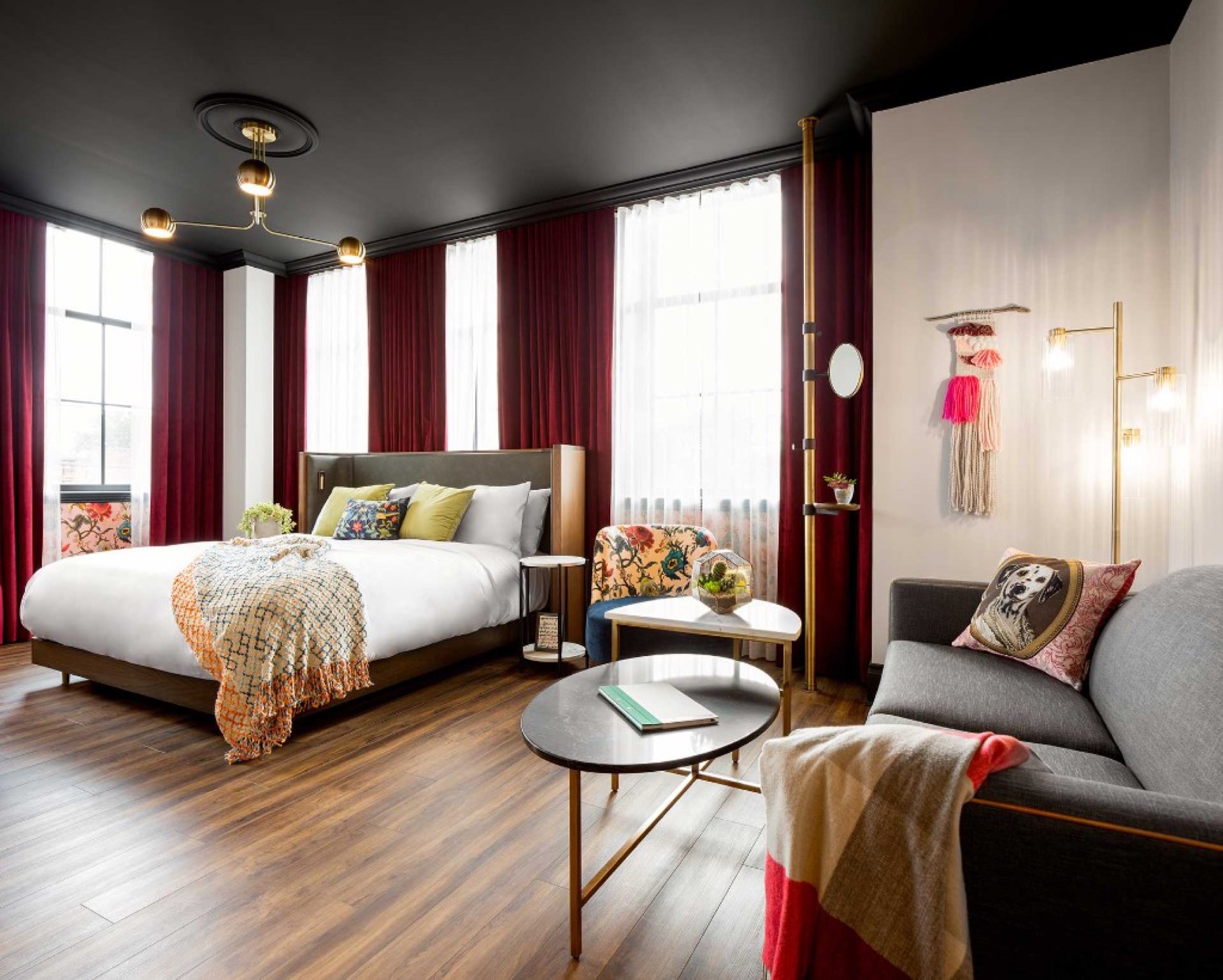 Boutique hotel king room with burgundy drapes, white bed linens, grey sofa with patterned pillows, floral armchair, marble coffee tables, and black ceiling with brass chandelier