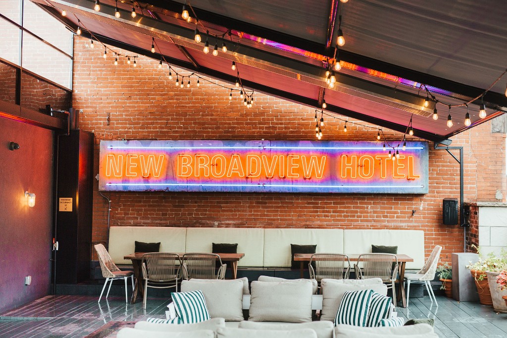 Rooftop lounge at The Broadview Hotel with New Broadview Hotel neon sign on brick, string lights, grey outdoor seating, and striped cushions