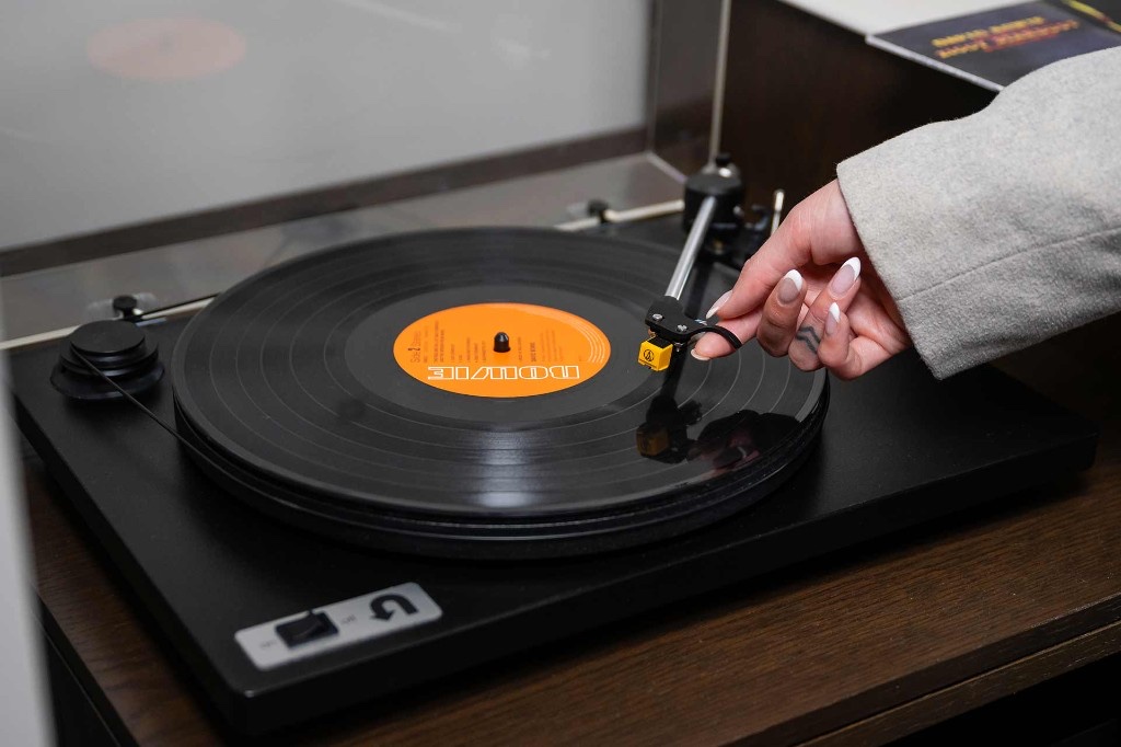 Hand lowering a tonearm onto a spinning vinyl record on a black in-room turntable, orange label on the disc
