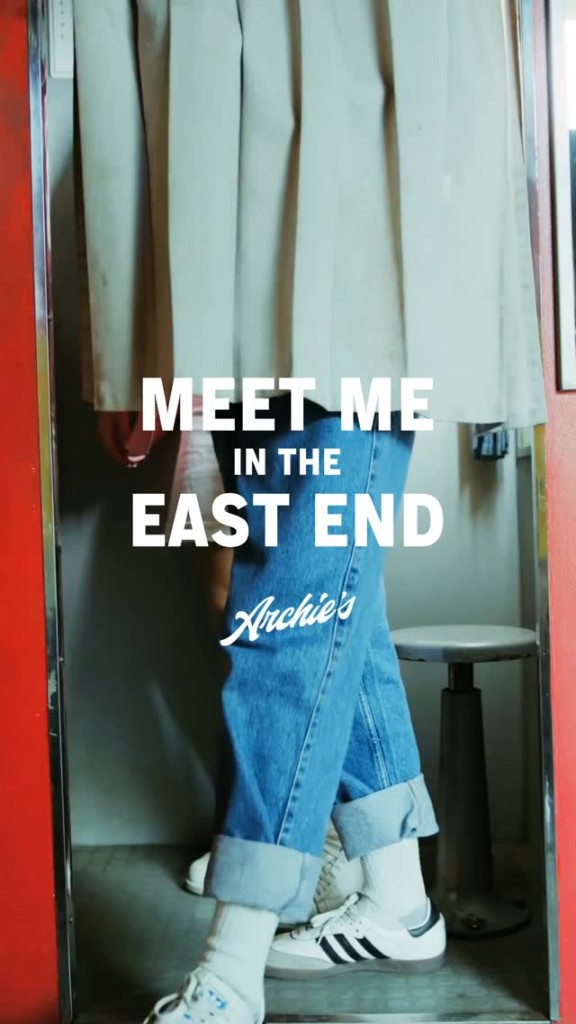 Legs in cuffed jeans and white sneakers in a photo booth with red frame; white text Meet Me in the East End and Archie's script